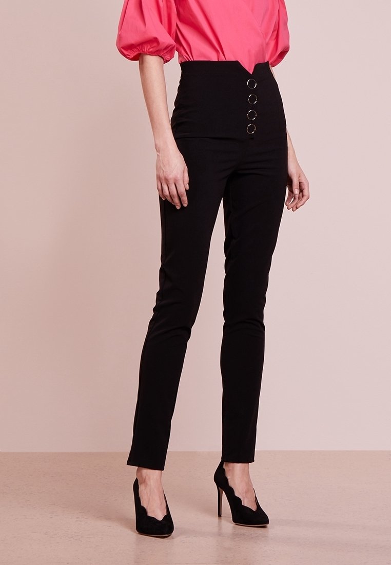 Femme portant un pantalon noir taille haute avec quatre boutons devant, associé à un chemisier rose à manches bouffantes et des chaussures noires à talons hauts.