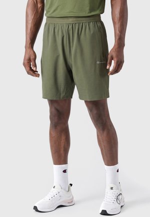 Mannelijk model draagt olijfgroene sportshorts, witte sokken met het Champion-logo en witte Champion sneakers, staand tegen een effen achtergrond.