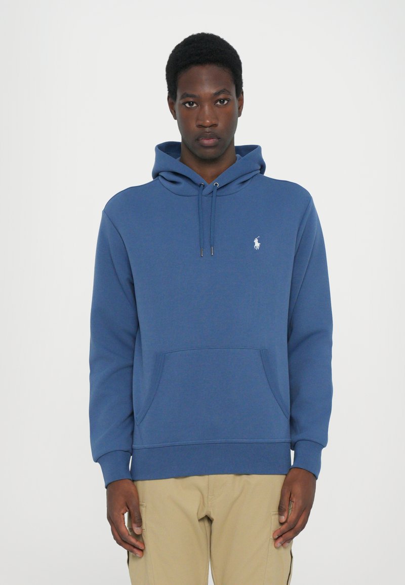 Mėlynas gobtuvas su raišteliu, priekinė kišenė ir išsiuvinėtas logotipas ant krūtinės. Pagamintas iš minkštos, tekstūruotos medžiagos.