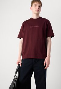 Burgundy cotton t-shirt featuring a round neckline and short sleeves with "Calvin Klein" logo in a subtle font, paired with dark denim pants.
