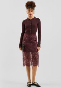 Burgunderfarbene gerippte Langarmbluse mit Kragen und Knöpfen, kombiniert mit einem Spitzen-Bleistiftrock mit gewelltem Saum; Accessoires: schwarze Clutch.