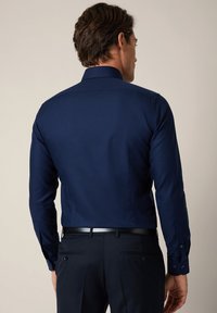 Granatowa koszula z długim rękawem, z usztywnionym kołnierzykiem, gładkim materiałem i rękawami zapinanymi na guziki. Posiada dopasowany krój i brak widocznych wzorów.