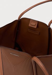 Bolsa de couro castanho com exterior texturizado, abertura superior que mostra um interior espaçoso, fecho de correr e logótipo em relevo de Karl Lagerfeld tanto dentro como fora.