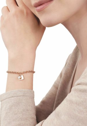 Mujer que lleva una pulsera de cadena en oro rosa con un dije en forma de corazón y una pequeña piedra preciosa clara, apoyando su mano cerca del rostro.