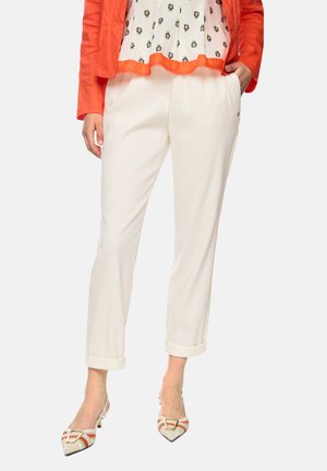 Woman wearing white rolled-up pants, orange patterned slingback heels, white blouse with orange trim, and orange jacket, hands in pockets.