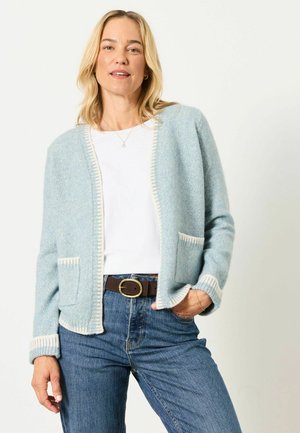 Woman wearing a light blue cardigan with white trim, white shirt, blue jeans, and brown belt, standing with one hand in pocket against white background.