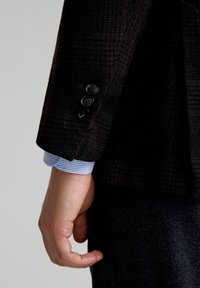 Manga de blazer de espiga oscura con tres botones negros; puño de camisa a rayas celeste visible debajo. Tejido de lana texturizada.