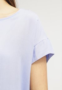 Světle modrý krátký rukáv top vyrobený z měkké tkaniny. Má volný střih a jemný detail srolovaného rukávu. Hladká textura a jednoduchý design.