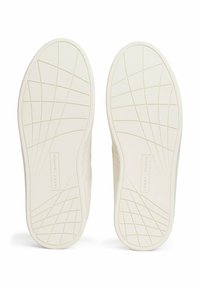 Białe podeszwie trampków z teksturowanym wzorem przyczepności oraz detalem loga. Materiał wydaje się gumowy, z gładkim wykończeniem i geometrycznymi wzorami.