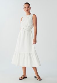 White, sleeveless maxi dress with a gathered waist and ruffled hem. Smooth texture, knee-length with a flowing silhouette, paired with black sandals.