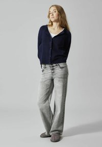 Marineblauer Strickcardigan über einem weißen Oberteil, kombiniert mit hellgrauen weit geschnittenen Jeans und grauen Hausschuhen. Einfaches Design, weicher Stoff und lässige Passform.