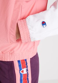 Chubasquero rosa con mangas blancas, puños elásticos y un logo bordado. Combinado con pantalones morados con rayas verticales y logotipos.