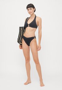 Black bikini with silver studs, halter neck design, keyhole detail at the bust, matching high-waisted bottoms, paired with a striped bag.