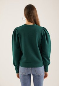 Groene, gestructureerde pullover met opgeblazen mouwen en een geribbelde zoom. Draagt lichte, strak zittende jeans, gezien van achteren.