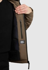 Brown outer jacket with a black padded interior; features a zip pocket and a logo patch; smooth fabric texture with a structured design.