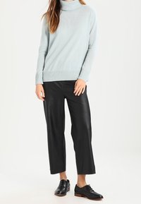 Frau, die einen hellblauen Rollkragenpullover, schwarze weite Hosen und schwarze Oxford-Schuhe trägt, steht vor einem einfarbigen Hintergrund.