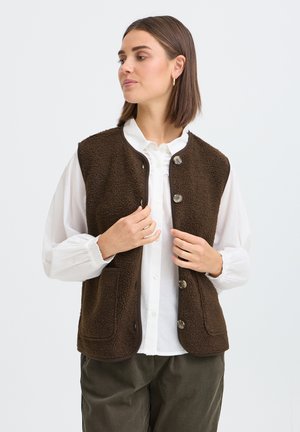 Femme portant un gilet en polaire marron foncé boutonné sur un chemisier blanc à manches bouffantes et un pantalon foncé, regardant sur le côté.