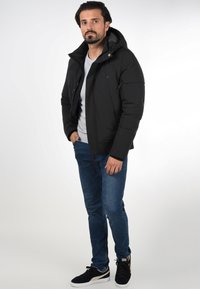 Sort polstret jakke med hætte, lynlås foran og sidelommer, båret over en grå skjorte og blå jeans, parret med sorte sneakers.