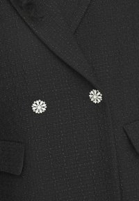 Veste noire texturée avec des broches florales doubles aux pétales blancs et aux strass. Couture détaillée le long des revers.