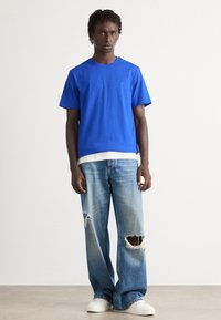 Camiseta de algodón azul de mangas cortas sobre una camisa blanca, combinada con unos vaqueros anchos de corte desgastado en azul claro y zapatillas blancas.