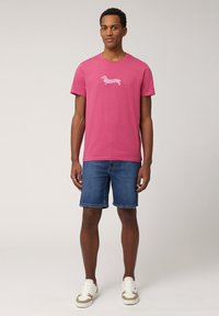 Camiseta de algodón rosa con un gráfico de un dachshund a rayas blancas en el pecho, combinada con pantalones cortos de mezclilla azul y zapatillas ligeras.
