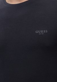 Μαύρο T-shirt με μικρό λογότυπο "GUESS EST. 1981" τυπωμένο στο αριστερό μέρος του στήθους, φορεμένο από ένα άτομο.