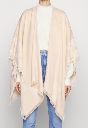 Personne portant un châle beige à franges sur un pull blanc et un jean bleu, debout devant un fond uni gris clair.
