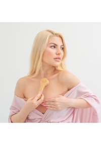 Szczotka do masażu z bambusa trzymana przez osobę w jasnoróżowym szlafroku, z gładką drewnianą rączką i miękkimi włoskami do pielęgnacji skóry.