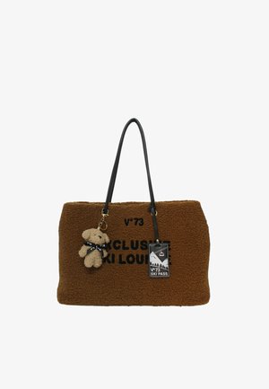 Bruine tote bag met textuur en zwarte handgrepen, zwarte tekst "Exclusieve Ski Lounge", een teddybeer sleutelhanger en een skipas tag.