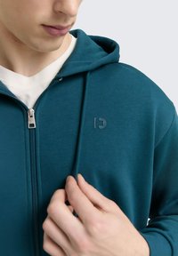 Sudadera con cremallera en color azul verdoso, con cordón, cremallera frontal y un logo en el pecho. Textura suave, usada sobre una camiseta de color claro.