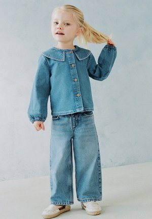 Jeune fille aux cheveux blonds portant une veste en jean bleue et un jean à jambes larges, tenant sa queue de cheval tout en se tenant contre un mur pâle.