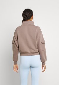 Brązowa krótka bluza z wysokim kołnierzem i detalem bocznej kieszeni, zestawiona z jasnoniebieskimi legginsami. Miękka faktura i luźny krój.