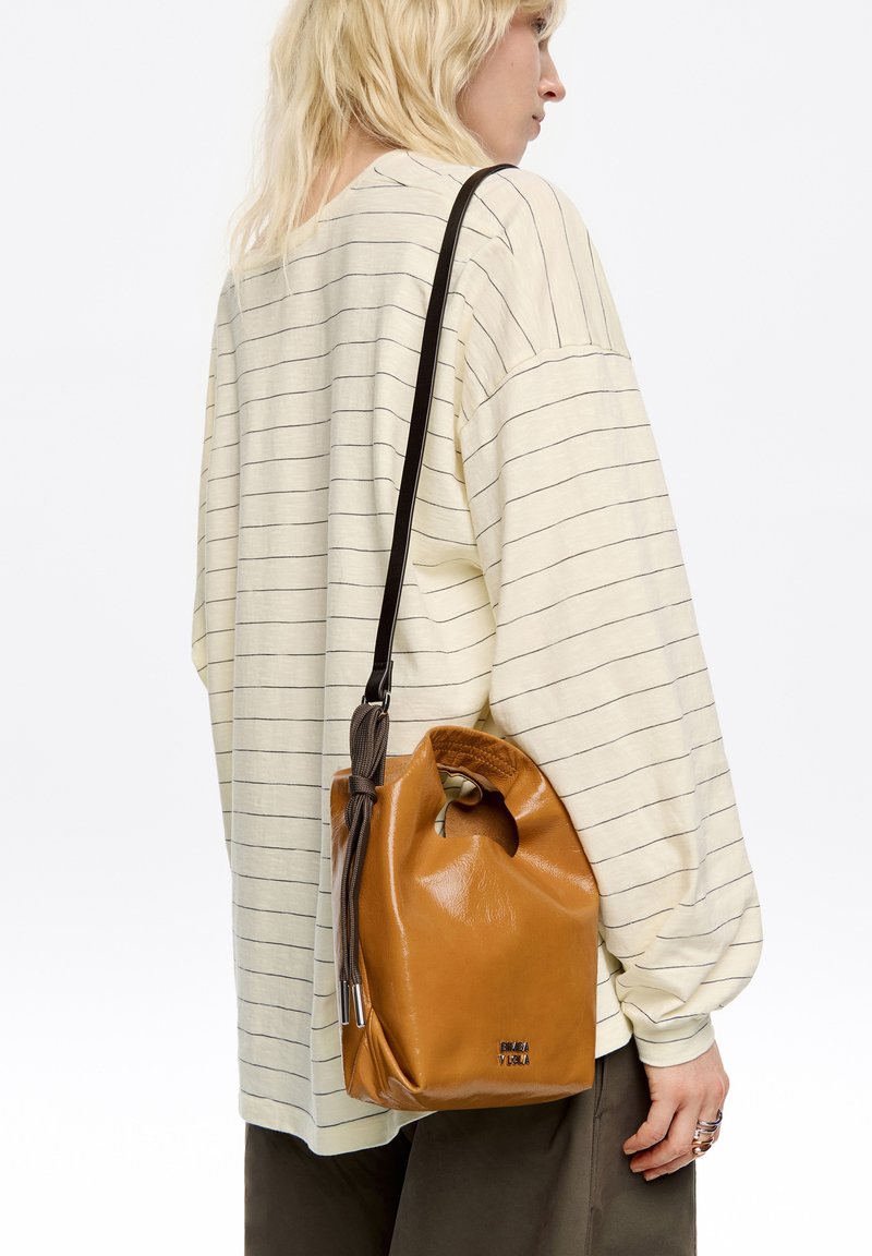 Skórzana torba w kolorze tan z zaokrąglonym dnem, z uchwytem do ręki i paskiem na ramię. Materiał ma gładką strukturę o delikatnym połysku.