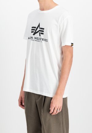 Mężczyzna w białej koszulce Alpa Industries z czarnym logo i napisem, w luźnych brązowych spodniach, na jednolitym białym tle.