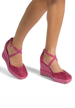 Des chaussures à talons compensés roses avec une base tissée texturée, ornées de clous brillants sur le bout et de sangles croisées à la cheville.