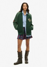 Veste bomber verte avec un logo, chemise bleu ciel à boutons, jupe à carreaux et bottes marron jusqu'aux genoux avec des clous.
