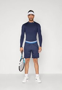 Σκούρο μπλε αθλητικό μπλουζάκι με μακριά μανίκια συνδυασμένο με αντίστοιχα σορτς, που έχει μπλε ζώνη, λευκό καπέλο τένις και κρατά μια ρακέτα τένις.