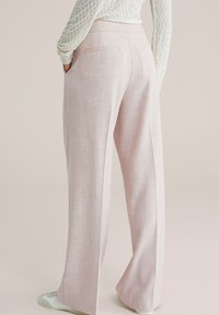 Osoba odjevena u svijetlo roza hlače širokih nogavica i bijelu teksturiranu majicu s dugim rukavima, ruke u džepovima, stoji na neutralnoj pozadini.