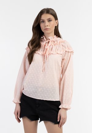 Blusa rosa chiaro con motivo a pois testurizzati, colletto a balze e colletto con lacci. Maniche lunghe con polsini arricciati. Indossata con pantaloni corti neri.