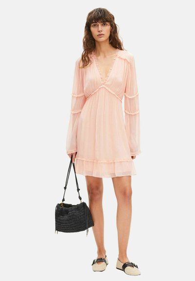 Mujer con vestido de manga larga color rosa claro y escote en V, sosteniendo un bolso negro tejido y usando zapatos beige sin cordones con correas negras.