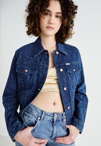 Chaqueta de denim en azul oscuro con patrón de olas embossado, botones de cobre y cuello clásico. Combinada con jeans de tiro alto en azul claro.