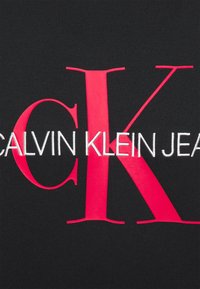 Il tessuto nero presenta "CALVIN KLEIN JEANS" in bianco e una grande "K" rossa. Il testo è in grassetto e il materiale ha una texture liscia.