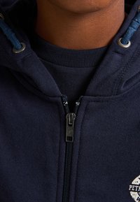 Sudadera con cremallera en color azul marino que presenta una cremallera plateada y cordones azules. El material es suave con una textura lisa; detalle del logo en blanco en la zona del pecho.