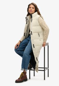 Gilet imbottito beige chiaro con colletto alto, dotato di hardware nero e molteplici tasche, indossato sopra un maglione di maglia e jeans blu.