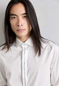 Camicia bianca con colletto strutturato, caratterizzata da un nastro verticale decorativo lungo la patta dei bottoni e una sottile ricamatura del logo.