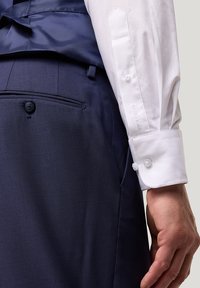 Navy-Hosen mit einer Vordertasche und Knopfverschluss, kombiniert mit einem strukturierten weißen Hemd mit Knopfmanschette und dezentem Branding.