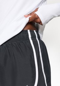 Mustad sportlikud lühikesed püksid valgete külgriidega, elastne värvel, sile tekstuur, koos valge pika varrukaga särgiga.