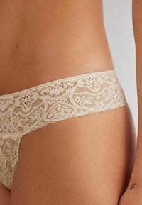 Beige kanten string met ingewikkelde bloemenpatronen langs de tailleband, kenmerkt zich door een zachte, doorschijnende textuur en een lage taille.