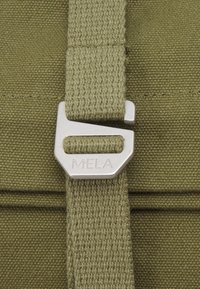 Green canvas bag detail featuring a metal buckle with “MELA” engraved, complemented by textured fabric and stitching accents.