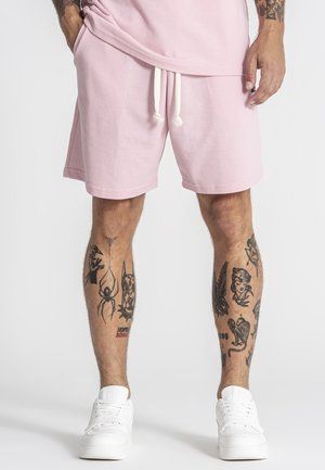 Man draagt lichtroze korte broek en witte sneakers, toont getatoeëerde onderbenen met verschillende zwarte inktontwerpen.